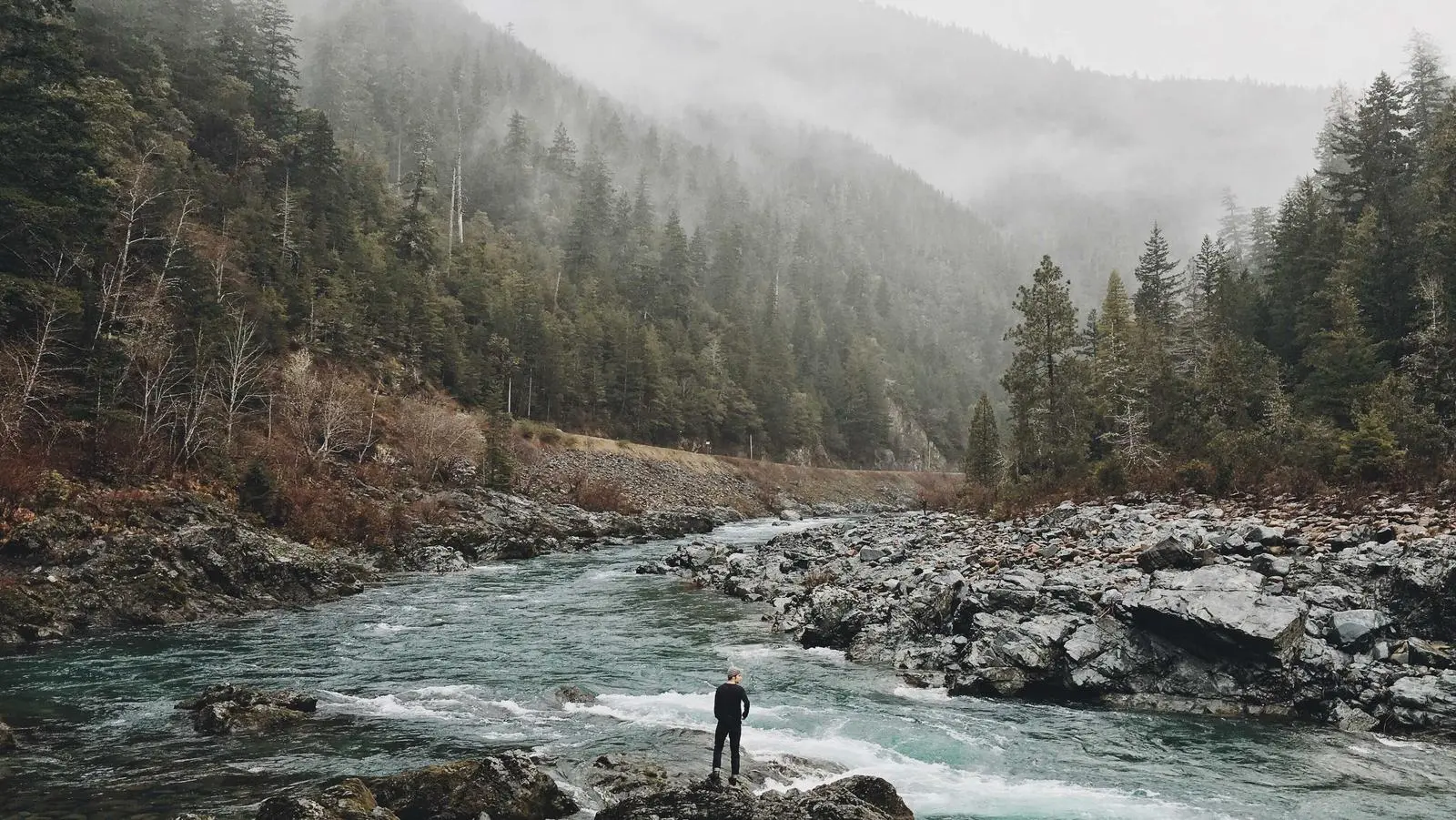 A person standing calmly by a river before misty mountains